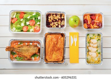 Fish, A Pie, Rice With Corn And Lean Chicken, Plums And Tangerines, An Apple, Tomato And Feta Salad, Hazelnuts, Dried Fruit And Plastic Cutlery On The Wooden Surface. Nutritional Meal Delivery.