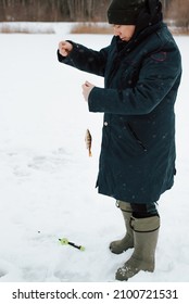 Fisherman Caught Fish On Winter Fishing Trip. Man Holding Small Perch On Fishing Line While Standing On Frozen Water, Outdoors.