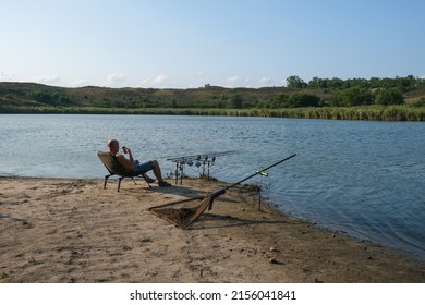 Fisherman with hot drink in mug waiting a bite with carp fishing equipment. Angler is fishing with carpfishing technique in summer evening, using rod pod, bite alarms, swinger, rods, landing net