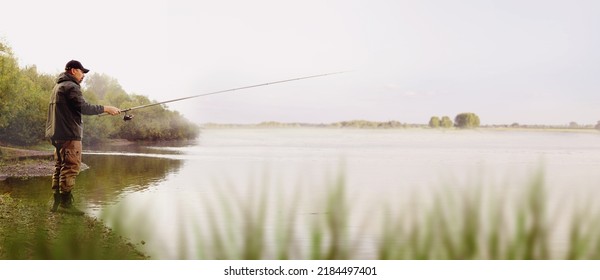 A Fisherman In A Jacket With A Fishing Rod Stands On The Bank Of The River. Fishing In The River. Fisherman With A Fishing Rod On The River Bank. Fishing, Spinning Reel, Fish. High Quality Photo