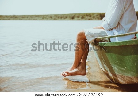 Fisherman relaxing on sunny day, cooling barefoot legs in lake water. Cut out image of unrecognizable male  sitting on the boat. Summer Holiday background. Copy space