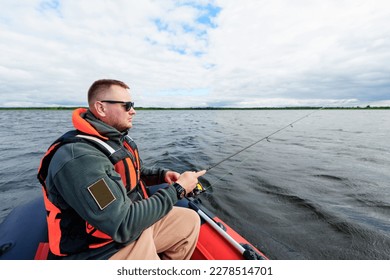 A fisherman sitting in a red boat with a fishing rod. Fishing on the lake in summer. A man in a life jacket