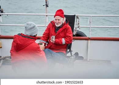 Fishermans Warming With Hot Beverage Before The Fishing. Hidden Camera View Of The Two Colleagues Wearing Warm Protective Clothes Is Smiling And Drinking Coffee In The Morning At The Boat Before The