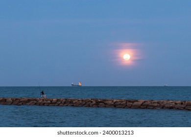 The fishermen with a fishing rod on a stone spit on the sea in the evening against the backdrop of a full orange moon and boat with lights