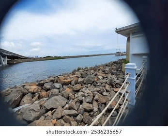 Fisheye view of coastal scene with rocky shore, calm water, distant bridge, and clear sky. - Powered by Shutterstock