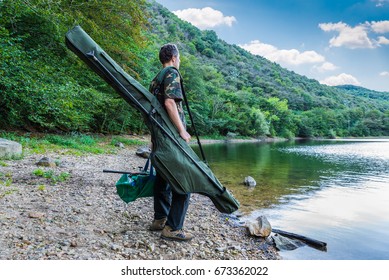 Fishing adventures, carp fishing. Fisherman on a lake shore with camouflage fishing gear, green bag and rod holdall