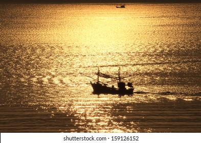 A Fishing Boat Is Coming Home In The Early Morning At Sunrise From The Work Of The Night In Prachuab, Thailand. It Crosses The The Golden Reflection Of The Rising Sun In The Water Of The Sea.