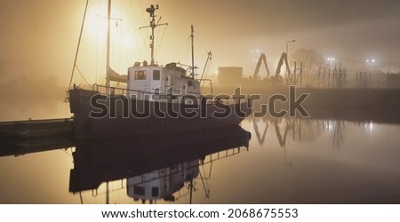 A fishing boat moored to a pier in a fog at night. Port cranes in the background. Yacht club illuminated by lanterns. Reflections on the water. Daugava river, Riga, Latvia