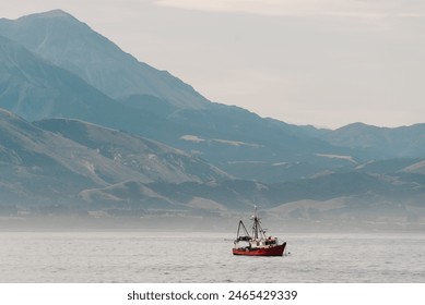 A fishing boat peacefully floats on the expansive surface of a vast body of water. The boat bobs gently as it navigates the calm waters with a magnificent mountain range in the misty background. - Powered by Shutterstock