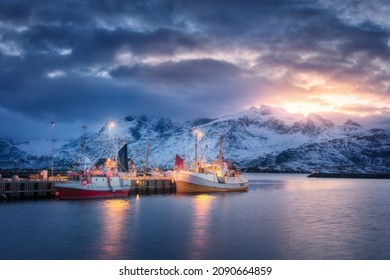 Fishing Boats On The Sea, Snowy Mountains, Colorful Sky With Clouds At Sunset In Lofoten Islands, Norway. Winter Landscape With Boats, Harbor, Lights, Houses With Illumination, Rocks In Snow. Nature