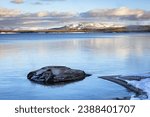 Fishing Cone Geyser Yellowstone Lake at sunset with reflections of snow-covered mountains, trees, shoreline and blue cloudy sky.