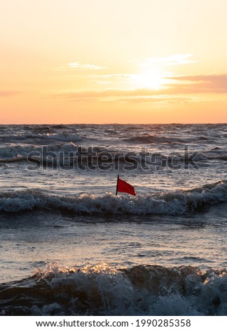 Fishing net location flag in the middle of the waves. Sunset on the background. Small red flag in the sea. Fishing location marked in  in the sea near the beach. Lobster pot location marked. Fishing
