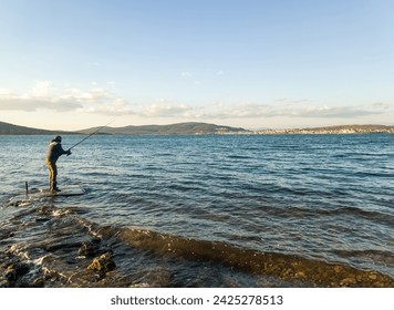 Fishing for pike, perch, carp. Fisherman with rod, spinning reel on sea or ocean. Man catching fish, pulling rod while fishing on sea, pond. Wild nature. The concept of rural getaway. Back view. 