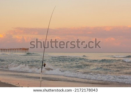 Fishing pole in surf on beach with pier in background 