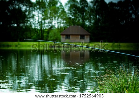 Fishing rod on the water lake with country house from wooden logs with green trees and grass on blurred background. misty cloudy weather. outdoors rest and relax concept