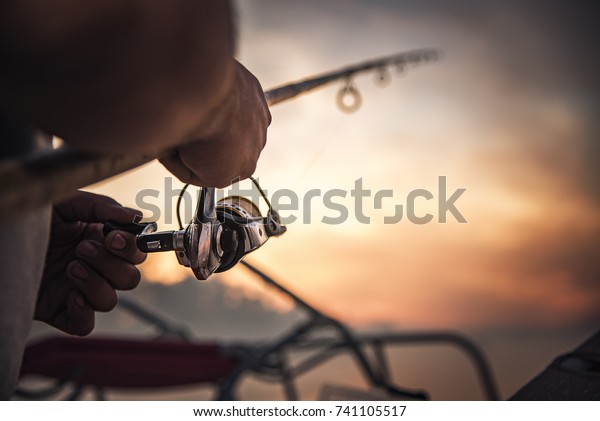 Fishing rod wheel closeup, man fishing with a\
beautiful sunrise behind\
him