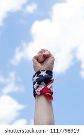 Fist With American USA Flag Tied On Wrist On Blue Sky Background. Struggle, Revolution And Fight Concept. Independence Day In USA.