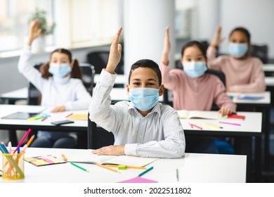 Fist Day Of School During Covid Pandemic Concept. Portrait Of Happy Hispanic Boy Sitting At Table In Classroom Wearing Disposable Medical Face Mask, Group Of Multicultural Small Pupils Raising Hands