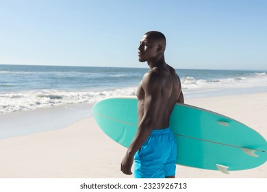 Fit african american man holding surfboard on sunny beach, looking away. Summer, healthy lifestyle, sport, hobbies, surfing and vacation, unaltered. - Powered by Shutterstock