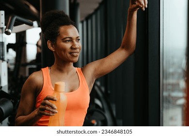 Fit African American woman standing next to the window and holding water bottle while smiling and looking into the distance. Fitness workout and hydration - Powered by Shutterstock