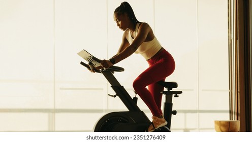 Fit African woman working out on a digital exercise bike, engaging in cardio to maintain her health and wellbeing. Woman using smart exercise equipment in her home fitness routine. - Powered by Shutterstock