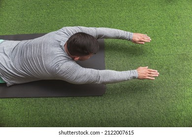 A Fit Asian Man Does Superman Back Extensions On A Black Mat. Isometric Exercise. Working Out And Training Lower Back Muscles At A Home Gym.