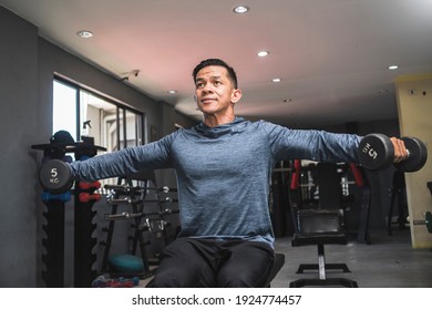 A fit asian man in a sweatshirt does seated side lateral raises. Shoulder workout and training. Open air gym setting. - Powered by Shutterstock