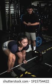 A fit asian woman does conventional barbell deadlifts while her instructor looks on.