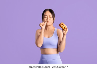 Fit asian woman in workout clothing, looking at tasty burger in her hand and licking her finger, contemplating diet choices against purple backdrop