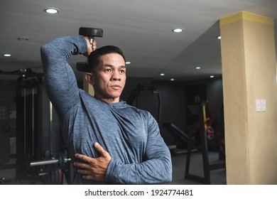 A fit athletic asian guy in a sweatshirt does seated overhead dumbbell one arm tricep extensions. Working out and training triceps. Open air gym setting.