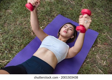 A Fit And Athletic Asian Woman Does Floor Dumbbell Flyes On A Yoga Mat On The Grass. Outdoor Chest Training Workout.