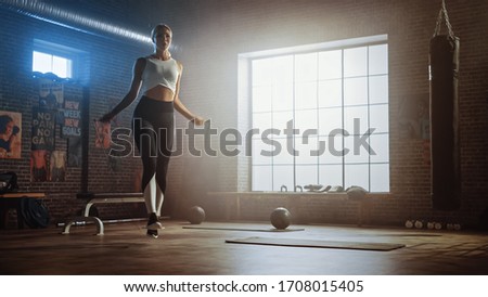 Fit Athletic Blond Woman Exercises with Jumping Rope in a Loft Style Industrial Gym. She's Concentrated on Her Intense Cross Fitness Training Program. Facility has Motivational Posters on the Wall.