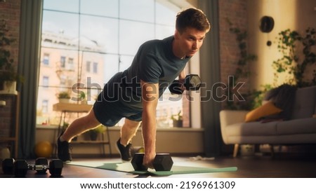 Fit Athletic Strong Young Man Performing Enduring Training in Plank Position, while Lifting Dumbbells During Morning Workout at Home in Cozy Bright Apartment. Concept of Healthy Lifestyle and Fitness.