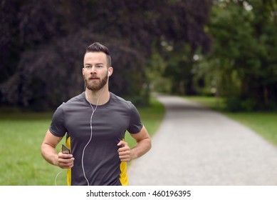 Fit Bearded Young Man Jogging Through A Park Listening To Music On His Mobile Phone, Upper Body Approaching The Camera With Copy Space