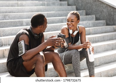 Fit Black Couple Taking Rest After Workout Outdoors, Sitting On Urban Stairs, Drinking Water And Coffee And Chatting, Free Space - Powered by Shutterstock