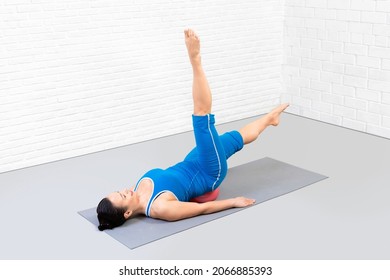 Fit caucasian woman in blue sportswear lying on back and does pilates drill with small fit ball under her lower back in fitness studio indoor.