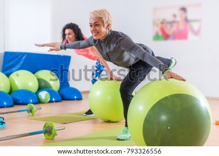 Fit Caucasian women doing crane exercise standing on one leg with their arms out to sides holding on to balance ball while training in fitness club