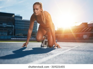 Fit And Confident Woman In Starting Position Ready For Running. Female Athlete About To Start A Sprint Looking Away. Bright Sunlight From Behind.