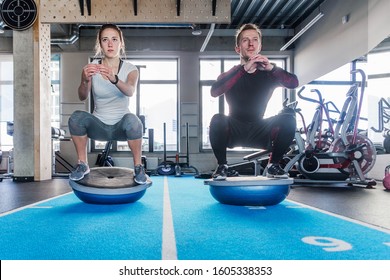 Fit Couple Working On Bosu Ball In Fitness Studio. Doing A Squat Exercise