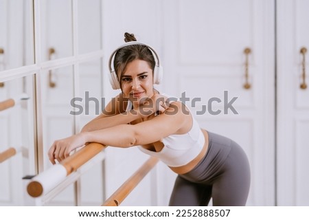 Fit European brunette girl in sportswear using headphones  makes stretching exercise at healthy club, leans on dance bar, looks at camera. Beautiful young fitness trainer ready for training. Fitness.