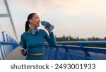 Fit European woman relaxing and drinking water on seaside promenade after running and training outdoors in the evening, panorama, copy space