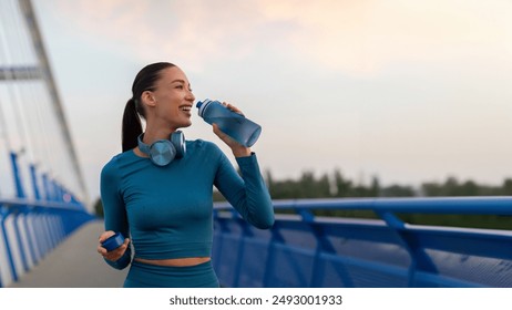 Fit European woman relaxing and drinking water on seaside promenade after running and training outdoors in the evening, panorama, copy space