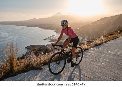 Fit female cyclist wearing cycling kit and helmet riding on the road on a gravel bike at sunset.Empty mountain road. Sports motivation image.Calpe town in Spain.