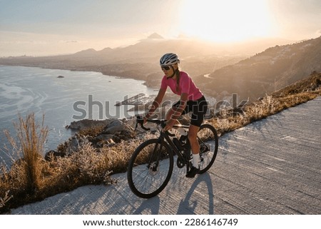 Fit female cyclist wearing cycling kit and helmet riding on the road on a gravel bike at sunset.Empty mountain road. Sports motivation image.Calpe town in Spain.