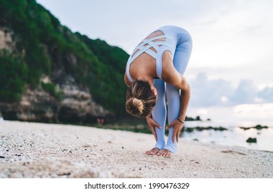 Fit female with hair bun in sports jumpsuit practicing yoga in Uttanasana pose on empty sandy beach with green rock on Bali