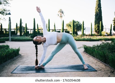 Fit female with long braid in sports top and leggins stretching in trikonasana triangle pose on black mat in park