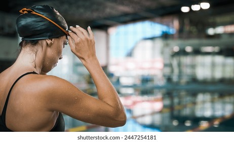 Fit female swimmer with a black swimming cap, standing looking at the pool and focusing on the coming race, putting on swimming goggles. - Powered by Shutterstock