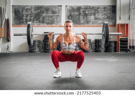 Fit female weightlifter in leggings and bra doing barbell back squat during functional workout in spacious gym in daytime