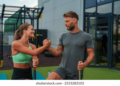 A fit friends in athletic wear are shaking hands and smiling at each other while exercising outdoors. They are in a gym setting with exercise equipment in the background. - Powered by Shutterstock