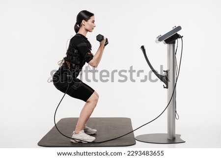 Fit girl does squats with dumbbells physical exercises in EMS suit that uses electrical impulses to stimulate muscles on white background. Sport training in electrical muscle stimulation suit
